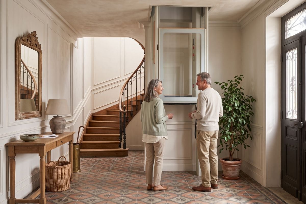 Couple souriant devant un ascenseur de maison Uplifts installé dans l’entrée d’une maison ancienne, près de l’escalier